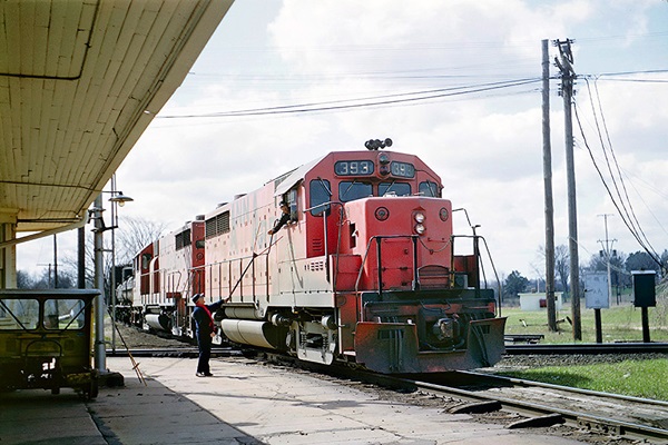 An engineer receives paper orders from a hoop in a red-painted locomotive leading a freight train.