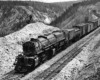 Mallet-type steam locomotive hauls a freight train on a rocky grade among mountain tops and thinning forest.