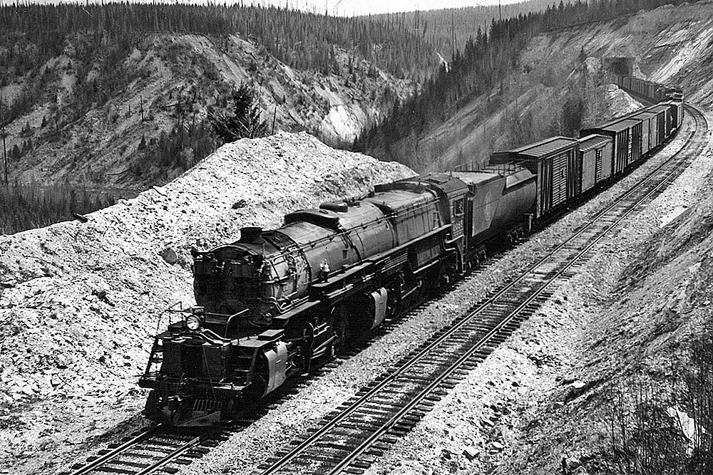 Mallet-type steam locomotive hauls a freight train on a rocky grade among mountain tops and thinning forest.