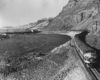 Diesel locomotive led freight train glides on an embankment above a river. A dam appears in the background of a treeless landscape.