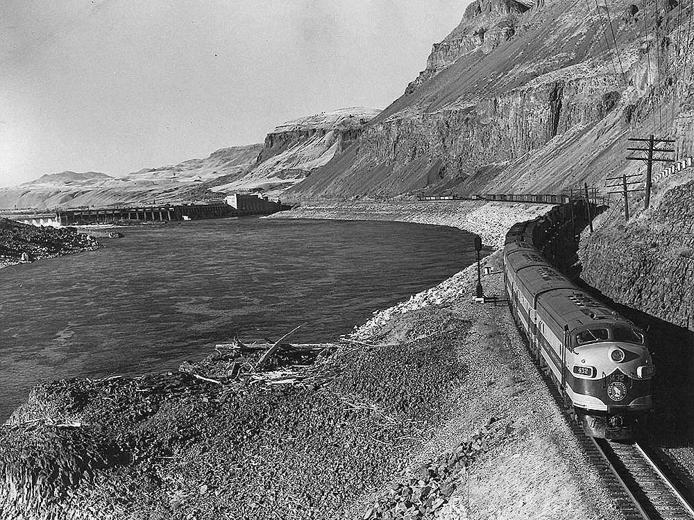 Diesel locomotive led freight train glides on an embankment above a river. A dam appears in the background of a treeless landscape.