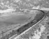 Two electric heavy freight motors pull a long freight train through a sparsely treed mountain scene.