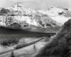 Cab-unit diesel locomotives lead a long passenger train on an s-curve at the foot of snow-capped mountains and forest.