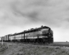Cab unit diesel locomotives lead a mail-express train through a prairie.