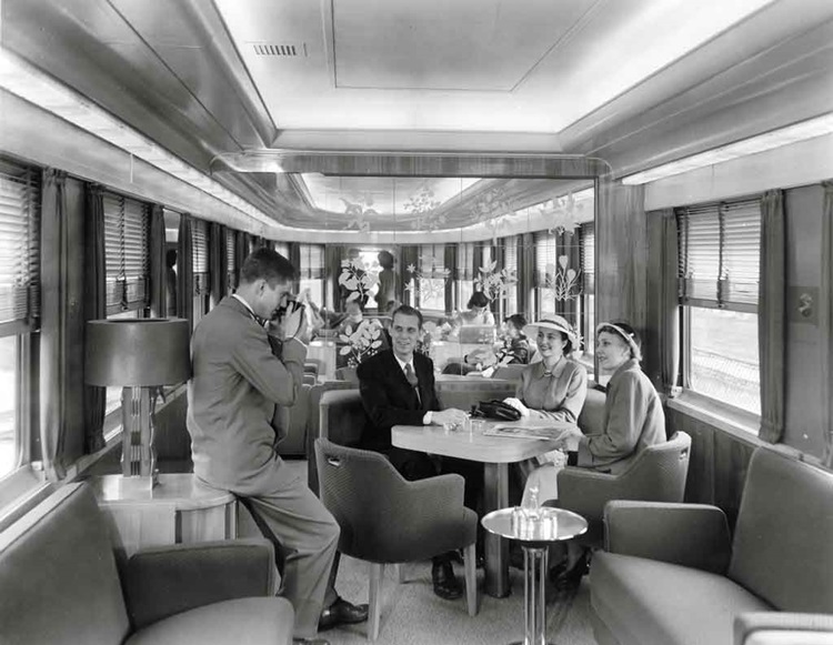 Man and two women seated at a passenger car table pose for a photograph taken by a second man.