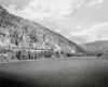 Passenger train in distance moves along a ledge above a wide river and below modest mountain tops.