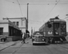 An interurban and an automobile appear stopped while a uniformed motorman walks to the cars with a stool in hand on a street.