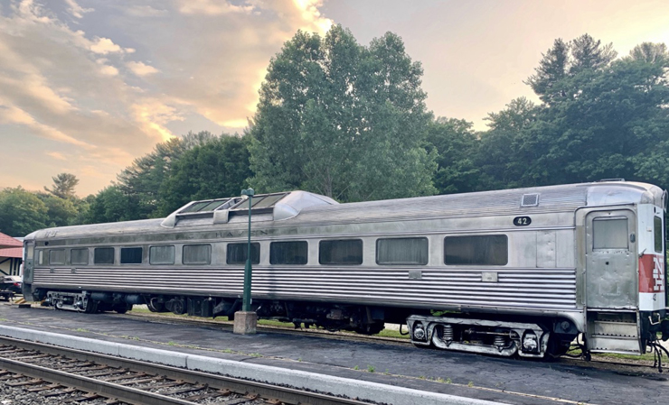 Silver-colored rail diesel car passenger unit with trees and low-angle sunlight sky in background.