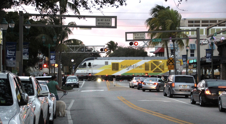 A yellow and white diesel locomotive leads a train through a busy intersection with cars, surrounded by palm trees.