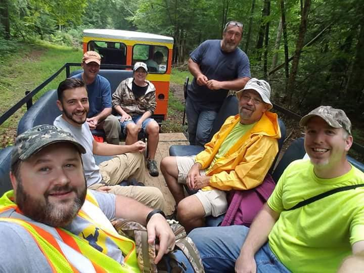 Seven men pose from an open car hauled by a speeder through a dense eastern hardwood forest.