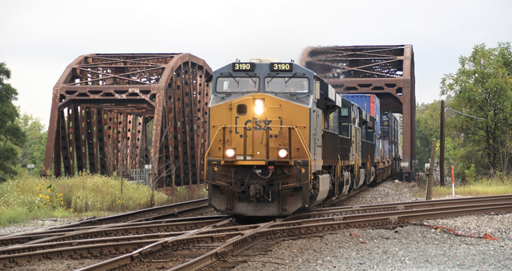 A blue-and-yellow painted locomotive leads an intermodal train through a truss bridge and over a double-diamond.