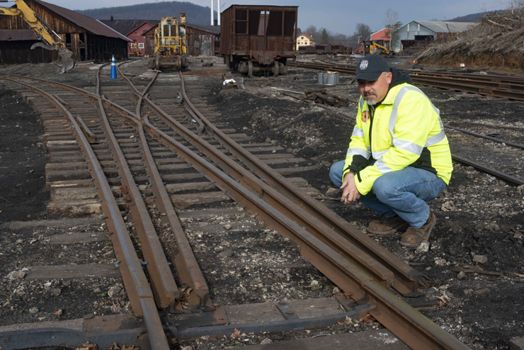 Man in high-visibility clothing squats near rust covered rails of a switch in a rail yard.