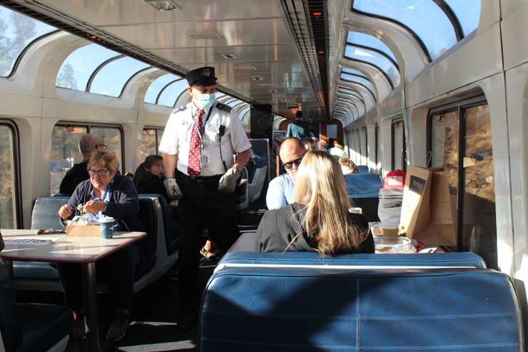 Masked Amtrak employee walking center aisle of a passenger car with broad arching windows.