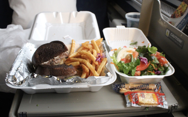 Sandwich and French fries in a styrofoam container beside a containered salad on a tray table.