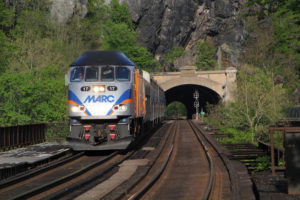 Blue and silver-painted diesel locomotive leads a train out of a tunnel in a forested scene.