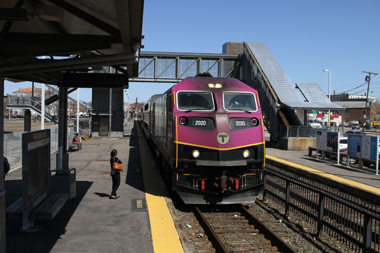 A purple painted locomotive heads a train surrounded by a passenger platform on a cloudless day.