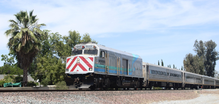 Silver painted train with red-striped nose of a diesel locomotive. Palm trees in the background.