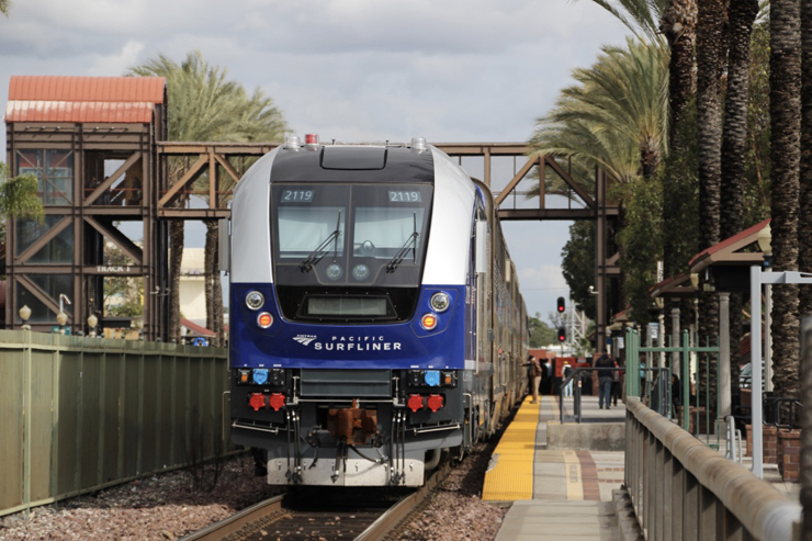 Blue and silver-painted locomotive  at a station platform surrounding by brown steel work and palm trees.