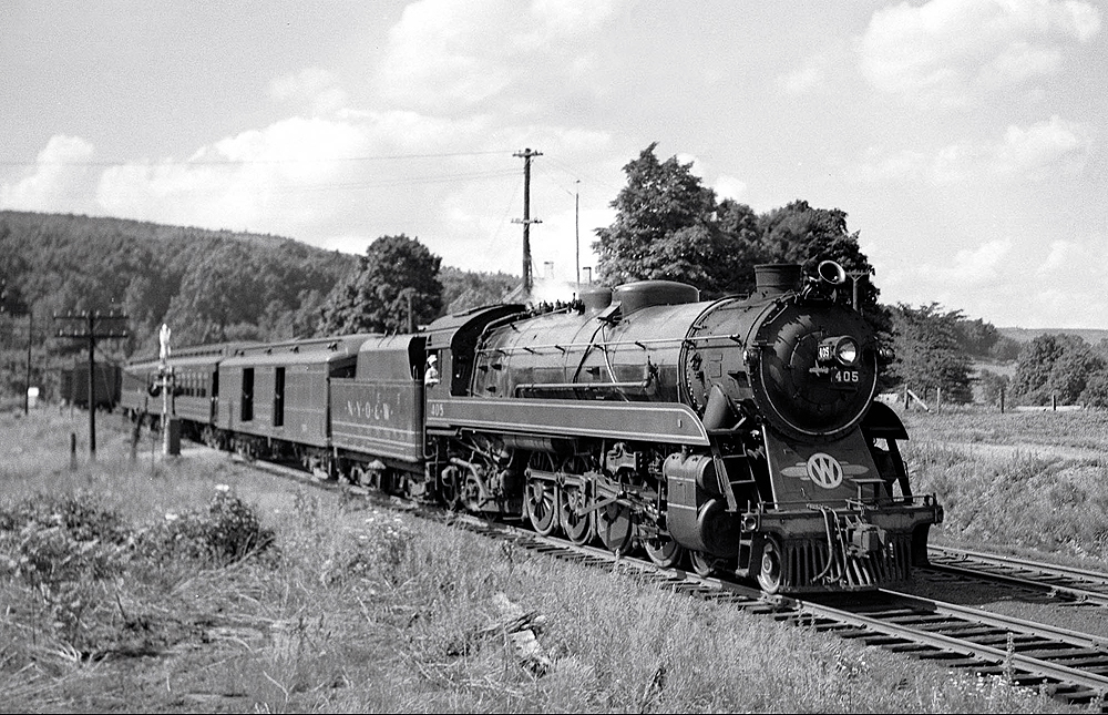 A steam locomotive hauls a passenger train through a forest-and-field covered valley.