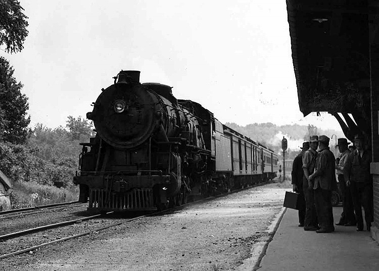 Steam locomotive leads a passenger train to a train station and waiting passengers.
