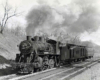 A steam locomotive leads a two-car passenger train through a cut.