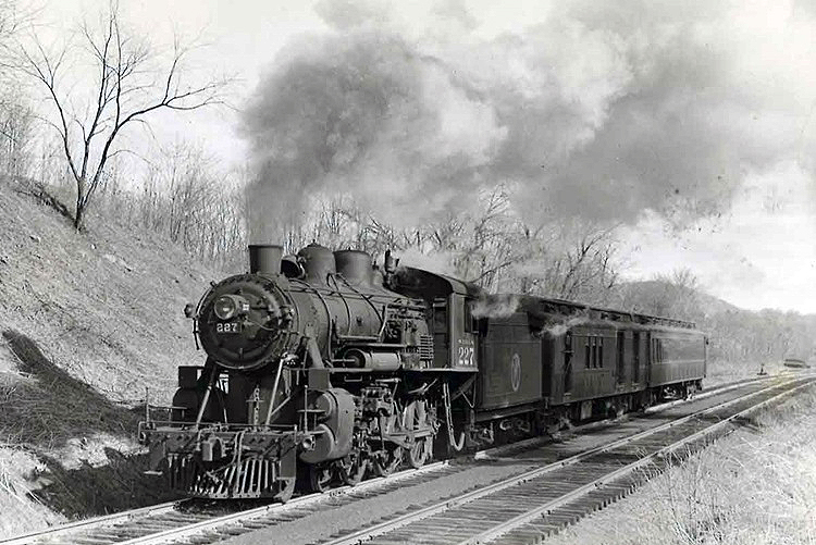 A steam locomotive leads a two-car passenger train through a cut.