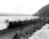 A steam locomotive leads a long train on a causeway between two water bodies at the foot of a mountain.