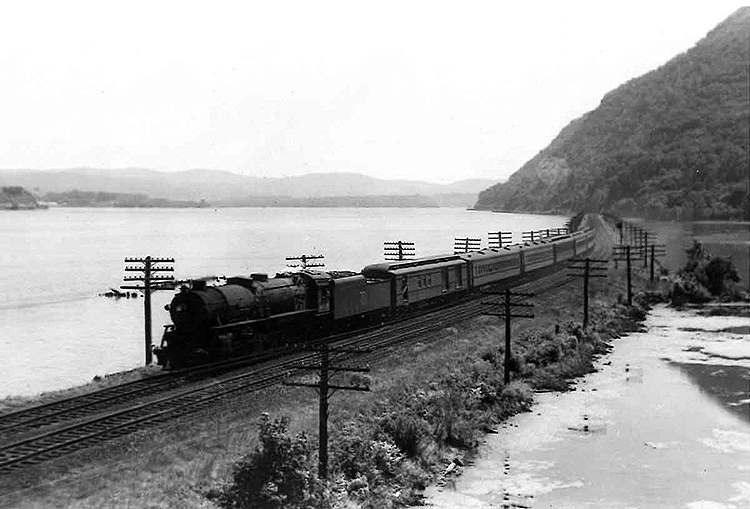 A steam locomotive leads a long train on a causeway between two water bodies at the foot of a mountain.