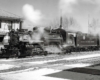 A narrow gauge steam locomotive leads a passenger train in front of a train station.