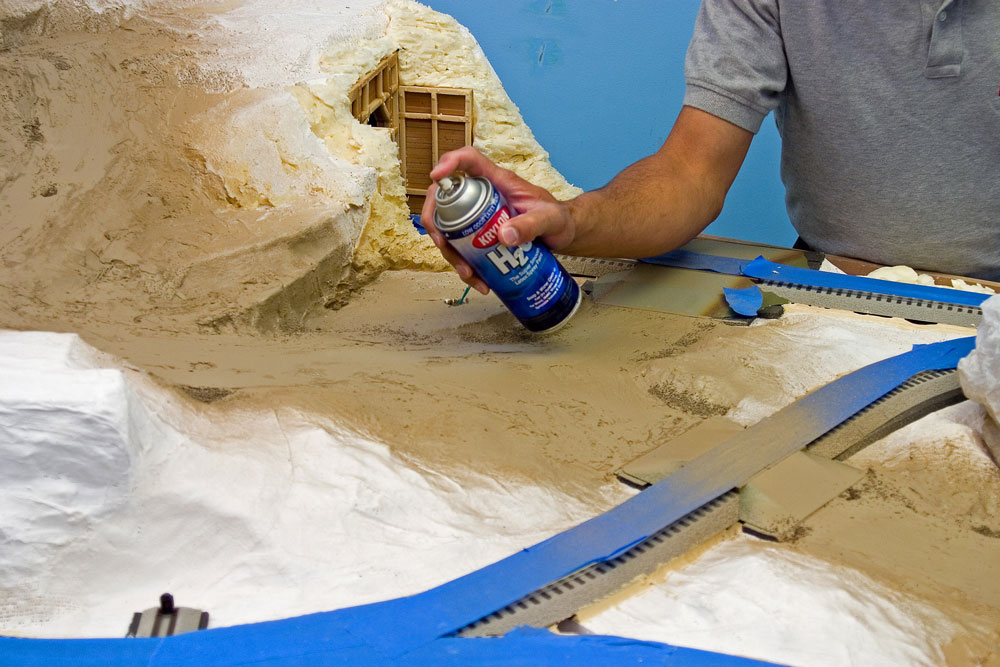 Man painting with water-based spray paint on model railroad layout.