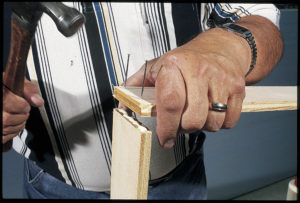 A man's hand holds a strip of plywood with two nails tapped in just above a perpendicular plywood strip coated with glue. He holds a hammer with his other hand.