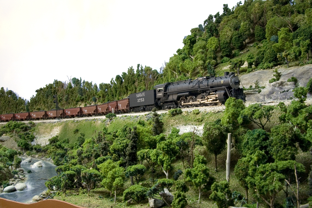 An HO Scale steam locomotive hauls uniform coal hoppers upgrade in a green mountain scene.