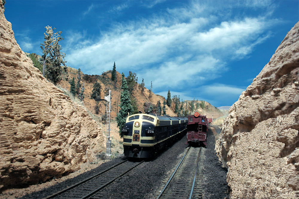 A blue-and-yellow Santa Fe diesel locomotive leads a train through an HO Scale stone walled gorge.