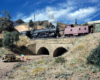 A Santa Fe steam locomotive helper pushes on a caboose upgrade through an arid-looking mountian landscape.