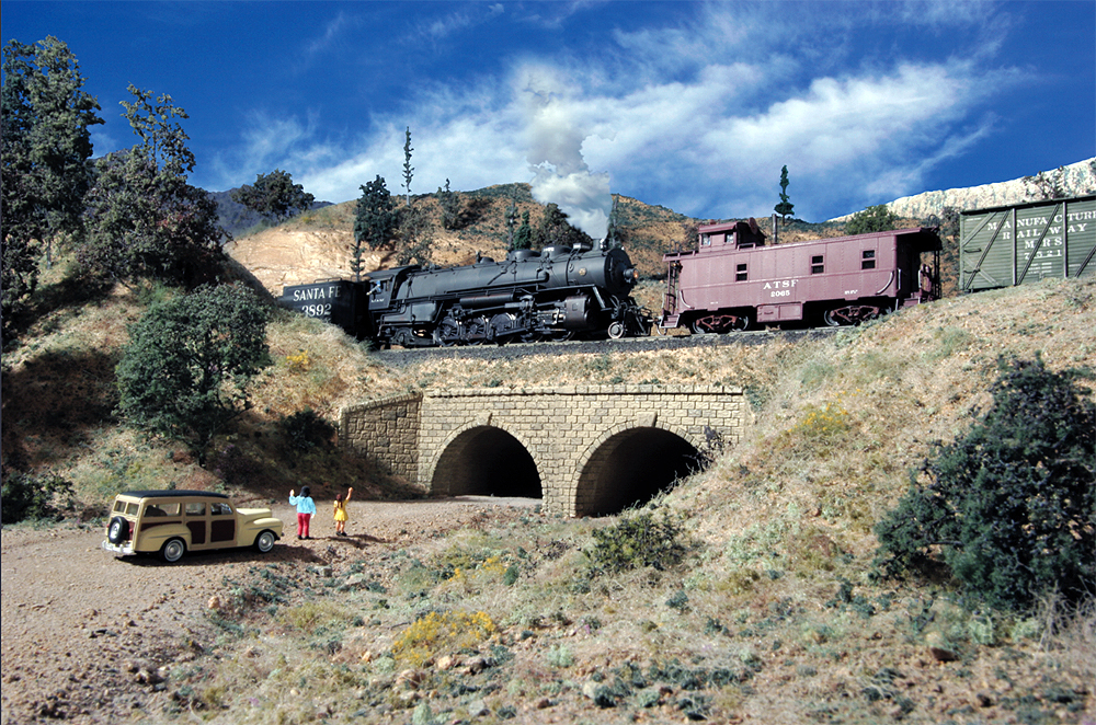 A Santa Fe steam locomotive helper pushes on a caboose upgrade through an arid-looking mountian landscape.