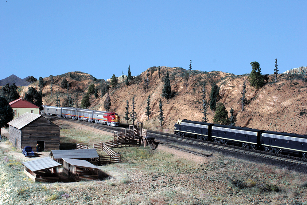 Red-and-silver Santa Fe passenger locomotives pull a train to meet with a blue-and-yellow Santa Fe freight locomotive-hauled train in an arid scene in front of a livestock loading area.