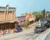 2-6-2 steam locomotives with freight train trundles down dirt-covered tracks in a small-town Western scene.