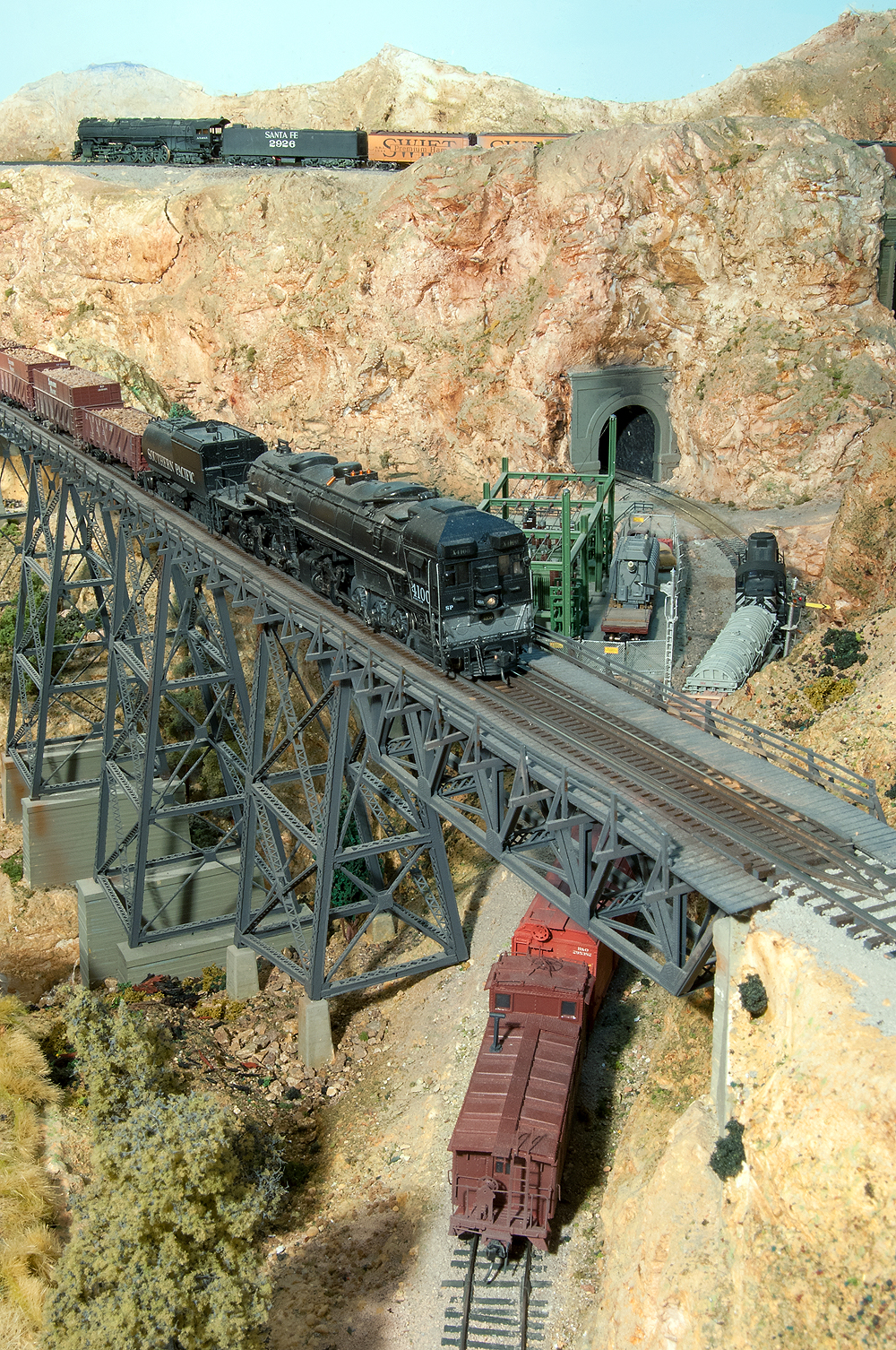 In the arid mountains along the Santa Fe rights-of-way, a Southern Pacific cab-forward steam locomotives operates on a trestle over a short Santa Fe diesel-powered freight train.