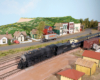 HO Scale Santa Fe steam and diesel locomotives team up to pull a train through a small town scene.