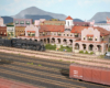 A 4-8-4 Santa Fe steam locomotive poses in front of a Harvey House hotel model amid a multi-tracked yard in a medium-sized city.