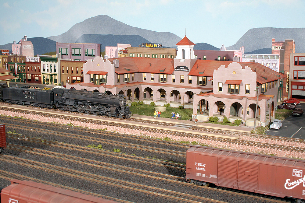 A 4-8-4 Santa Fe steam locomotive poses in front of a Harvey House hotel model amid a multi-tracked yard in a medium-sized city.