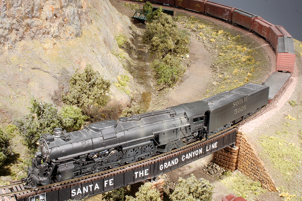 A heavy-duty steam-locomotive, likely a 2-10-4 hauls a train upgrade over a bridge on a Santa Fe-themed layout.