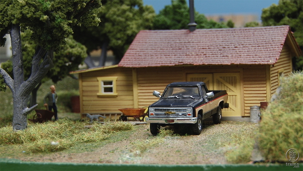 A scene on a model railroad that includes a house, truck, trees, and figure.