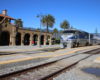 An Amtrak train paused at an adobe-style train station under clear skies with palm trees.