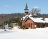 A blue-and-yellow diesel locomotive leads a freight train past a  gothic revival train station in snow.  