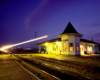 A light blur appears in front of an illuminated passenger train station in the evening.