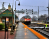 A stripped-nose locomotive pull a passenger train in rain by a small brick train station.