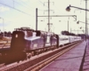 A streamlined electric locomotive hauling a passenger train beside a passenger platform.