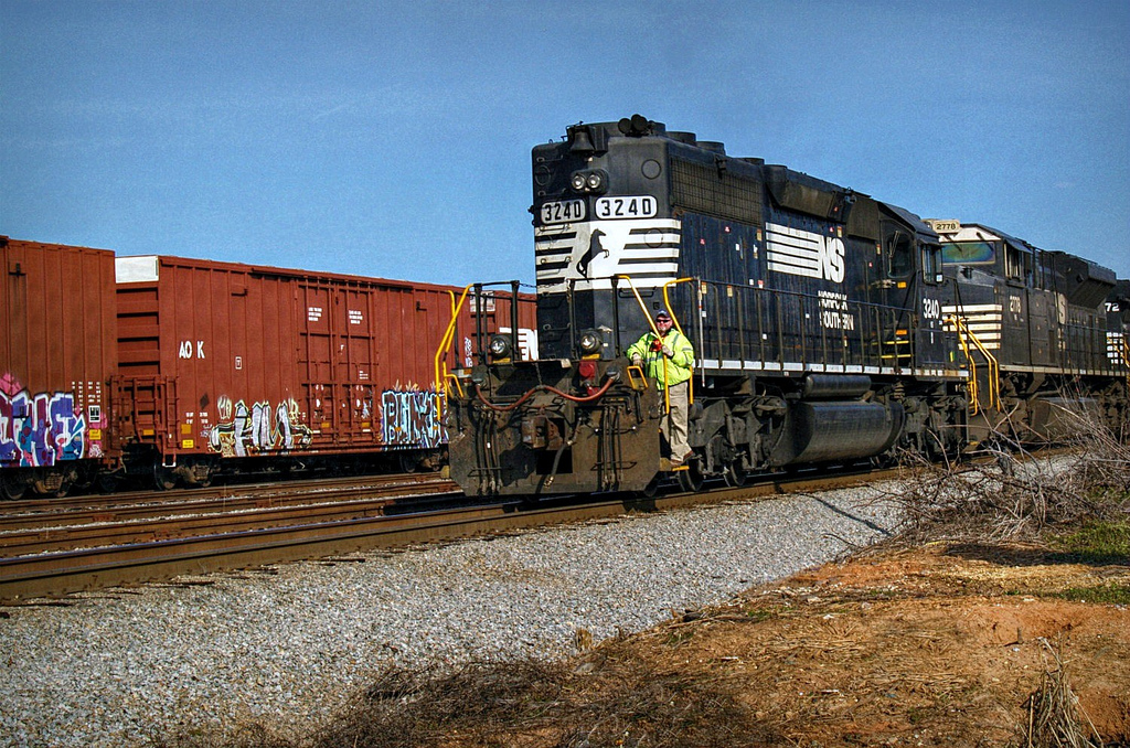 A railroad worker rides the steps of a black diesel locomotive in a yard.