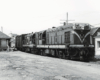 Two diesel electric locomotives at the head of a train amid a handful of age-worn buildings.